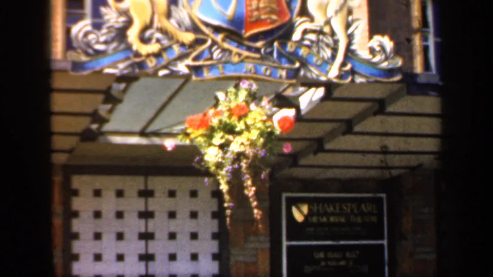 1949 England home-movie frame showing the crest and flowers above an entrance area with a Shakespeare Memorial Theatre sign visible at lower right.