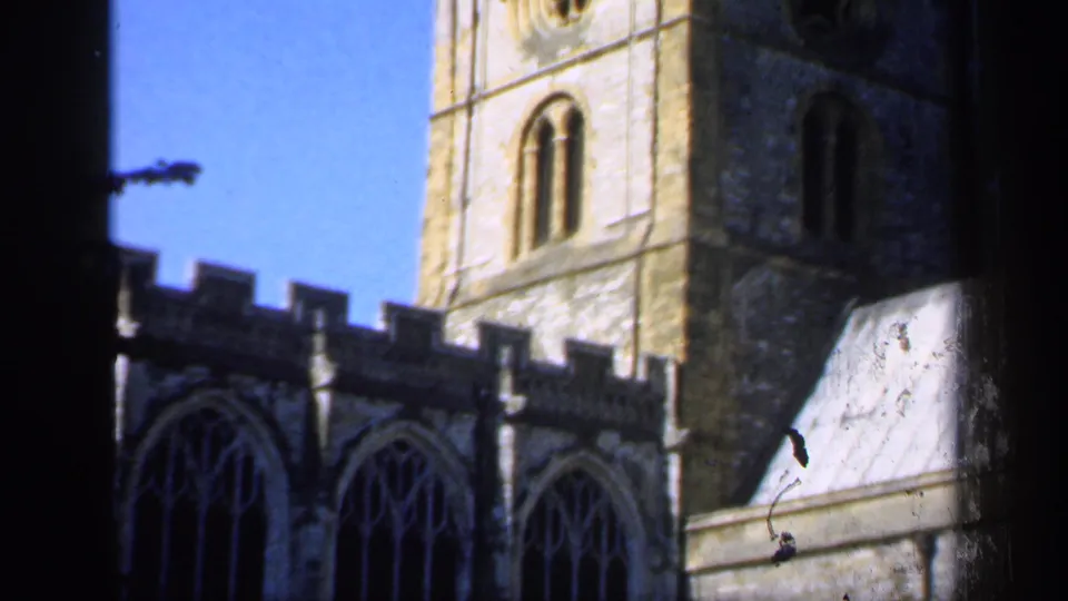 1949 England home-movie frame showing a stone church tower above Gothic windows and a pale adjoining roof, with bright sky behind.