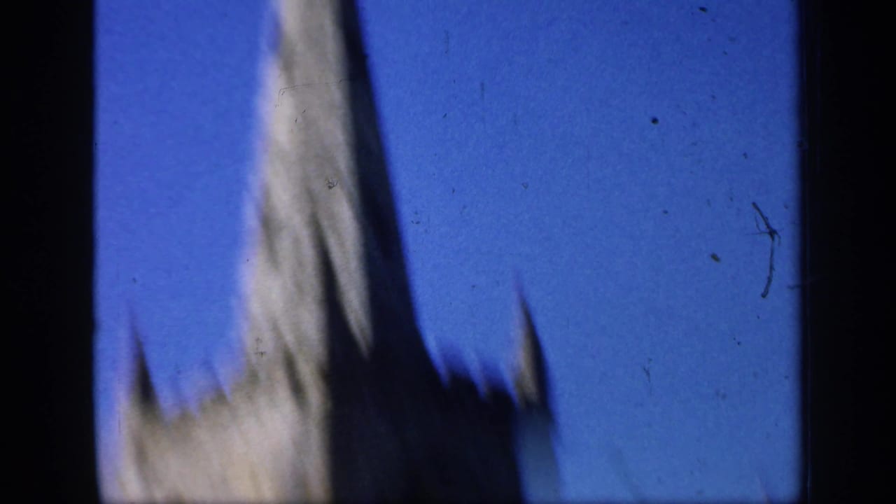 Soft 1949 England home-movie frame looking up at the upper part of a stone church spire against deep blue sky.