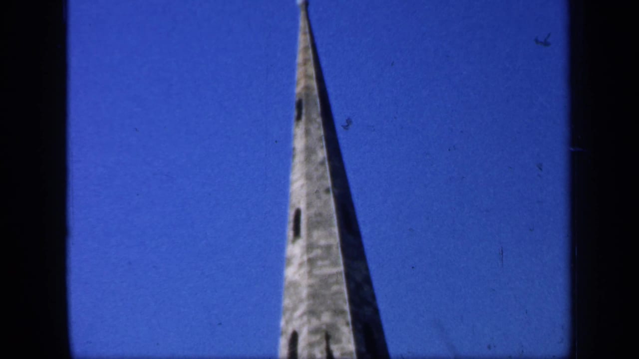 Tall English church spire filmed from below, with stonework and narrow openings visible against a blue sky.