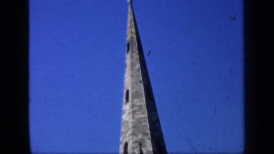 Tall English church spire filmed from below, with stonework and narrow openings visible against a blue sky.