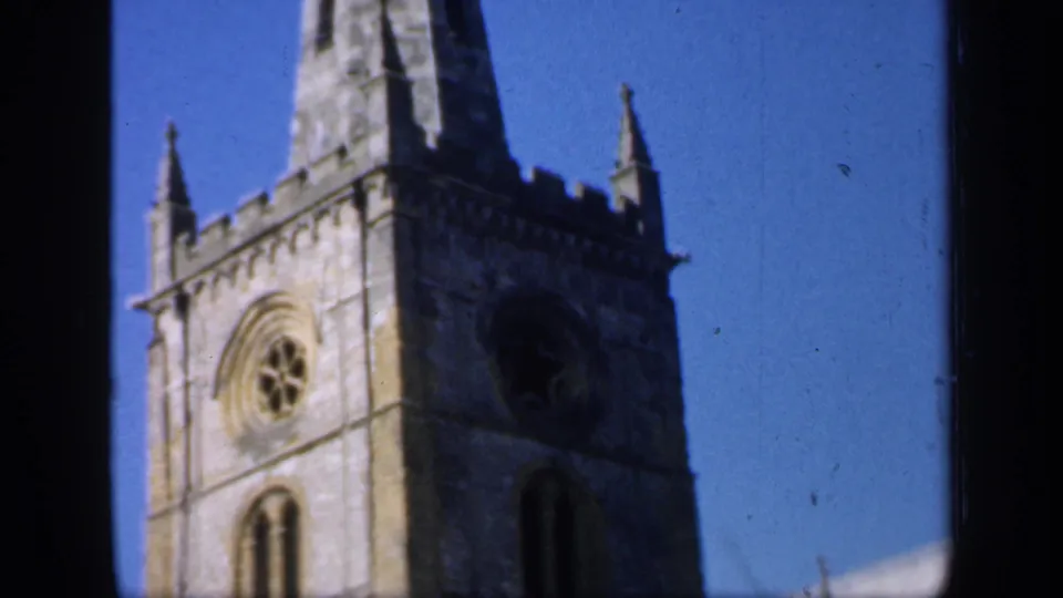 Handheld 8mm frame of an English stone church tower, showing the spire, crenellations, circular window, and arched opening.