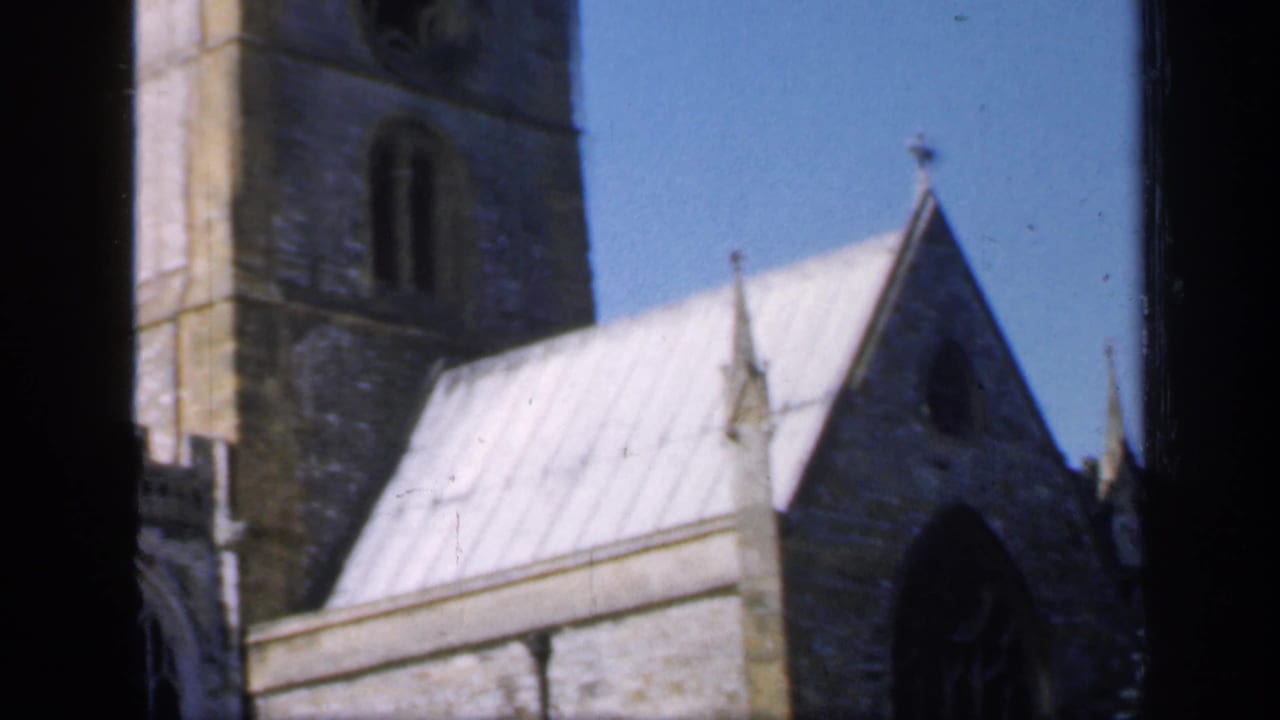 1949 England frame with a stone church tower behind a pale roof and pointed gable, divided by dark foreground window framing.