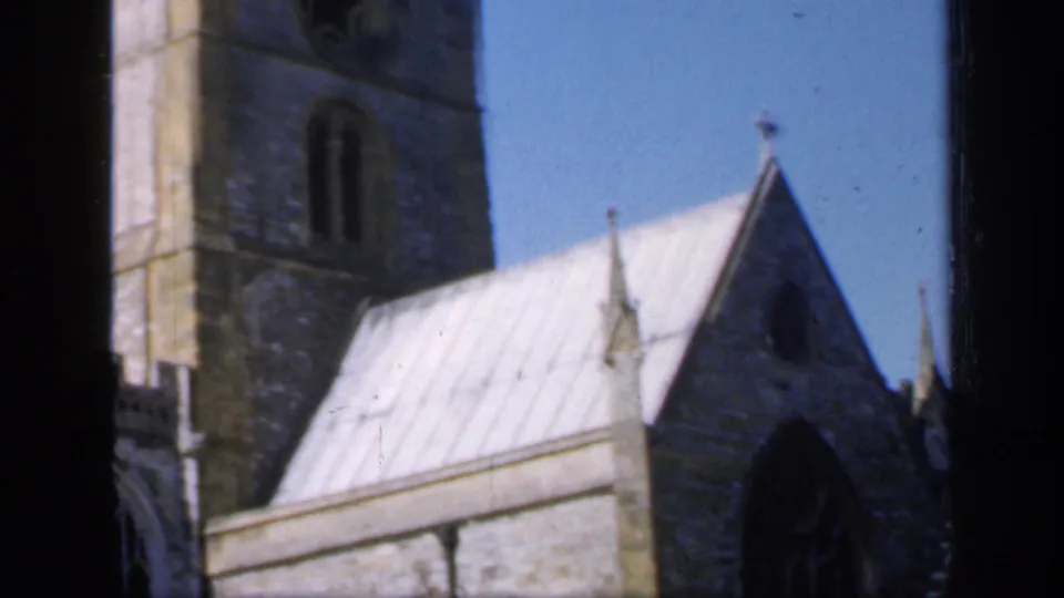 1949 England frame with a stone church tower behind a pale roof and pointed gable, divided by dark foreground window framing.