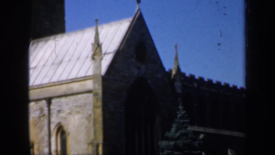 1949 England home-movie view of a stone church gable with a pale roof, arched window, dark foreground frame, and evergreen tree near the building.
