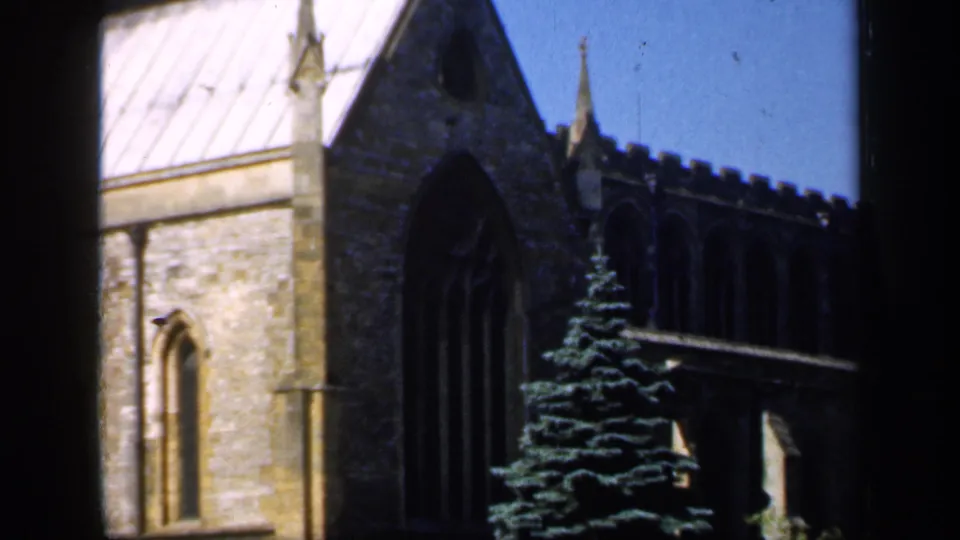 1949 England frame showing a stone church wall with tall Gothic arched window, pale roof, crenellated roofline, evergreen tree, and dark window mullions in front.