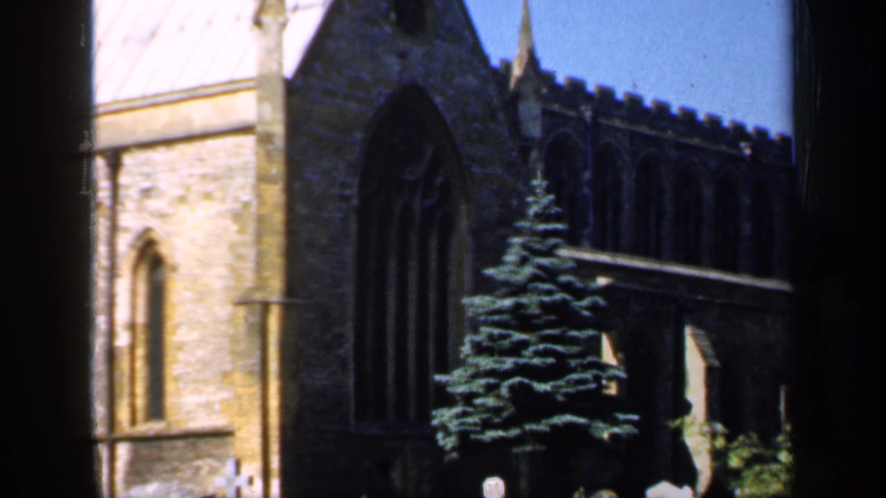 1949 England 8mm frame showing a stone church with Gothic windows, an evergreen tree, and churchyard headstones viewed through a window frame.