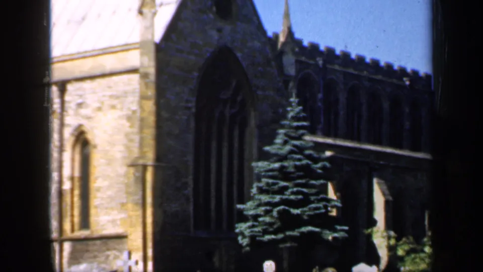 1949 England 8mm frame showing a stone church with Gothic windows, an evergreen tree, and churchyard headstones viewed through a window frame.
