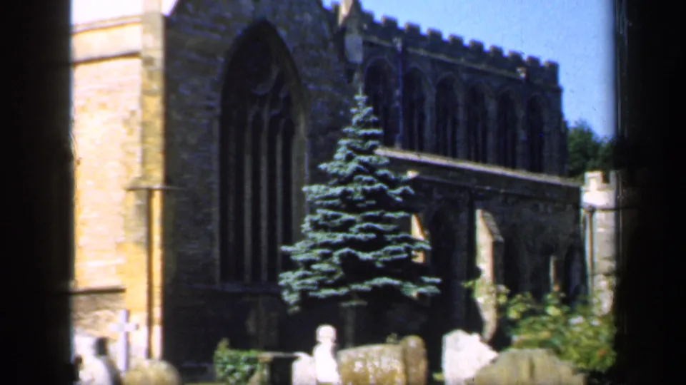 1949 England home-movie frame of a Gothic stone church, tall evergreen, and weathered churchyard headstones under bright daylight.