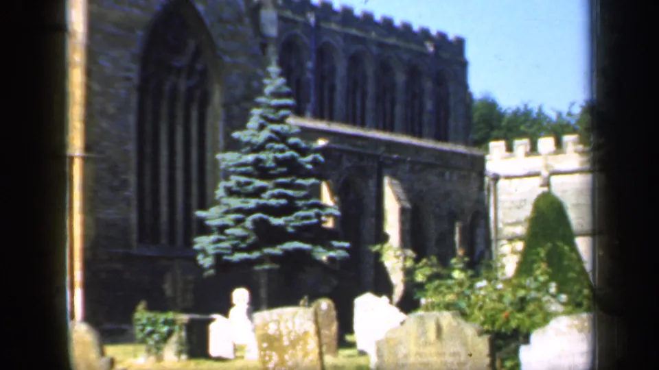 1949 England 8mm view of a churchyard with a prominent evergreen, Gothic church windows, shrubs, and several headstones.