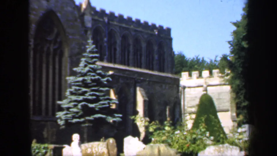 1949 England frame showing an evergreen tree in front of a Gothic stone church, with headstones and garden greenery in the churchyard.