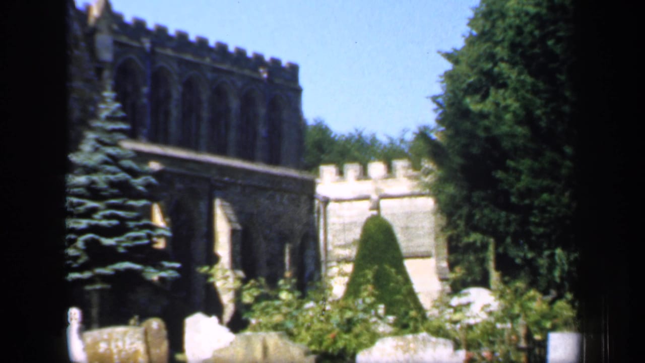 1949 England home-movie frame of a churchyard with crenellated stonework, Gothic arches, grave markers, clipped shrub, and trees.