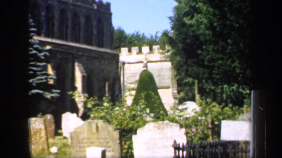 1949 England 8mm view of tombstones and an iron fence in front of a historic stone churchyard with clipped shrubs and trees.