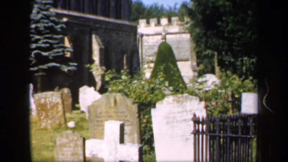 1949 England home-movie frame showing larger foreground tombstones, a black iron fence, Gothic church stonework, and green churchyard planting.