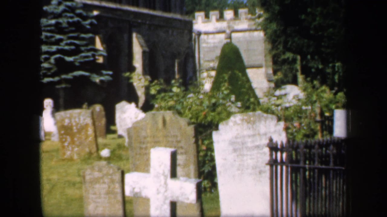 1949 England 8mm view of a sunlit churchyard with weathered gravestones, a white cross, clipped shrubs, and stone church architecture.