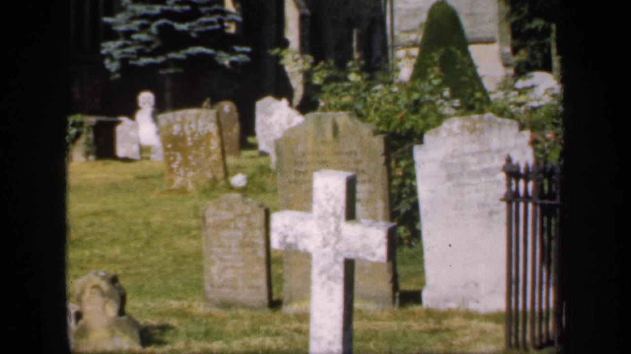 1949 England home-movie frame showing churchyard graves, a prominent white cross, iron fencing, grass, shrubs, and stone church fabric.