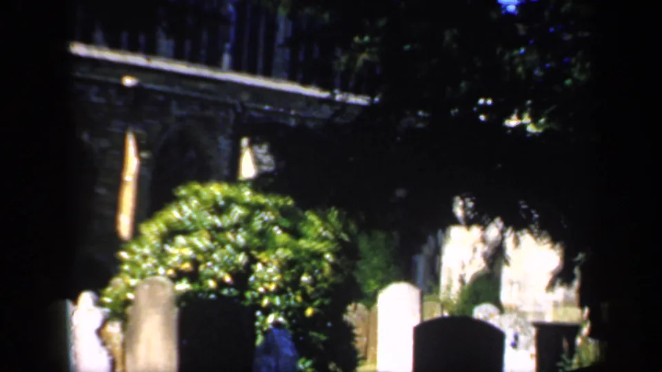 1949 England 8mm churchyard shot with deep shade, sunlit greenery, headstones, church stonework, and a small pale grave marker or figure.