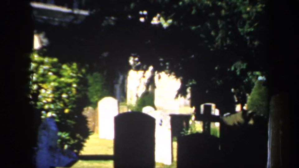 1949 England 8mm frame of a shadowed churchyard, with bright headstones, a dark foreground marker, trees, shrubs, and a cross-shaped grave marker.
