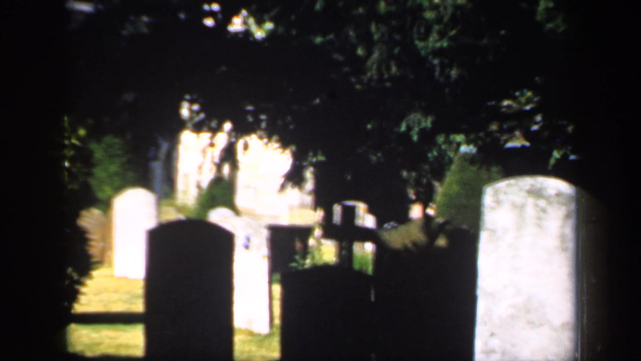 1949 England 8mm frame showing headstones in a shaded cemetery with bright patches of grass and trees.