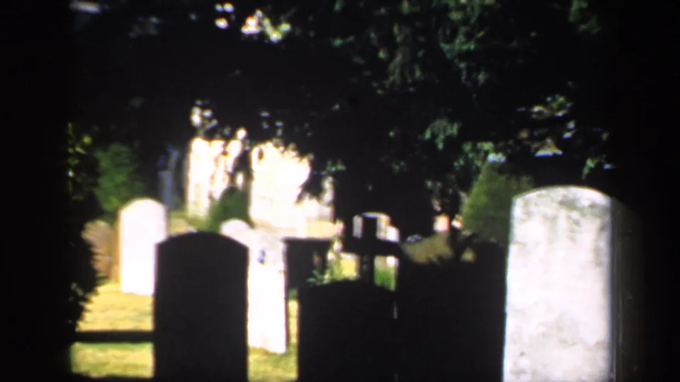1949 England 8mm frame showing headstones in a shaded cemetery with bright patches of grass and trees.