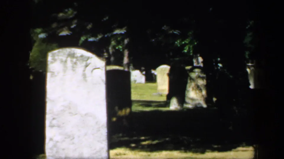 1949 England 8mm still with a large foreground headstone and scattered cemetery markers receding into shade.