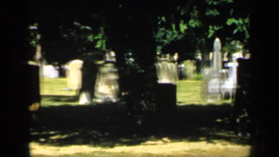 1949 England cemetery frame showing multiple headstones across a grassy burial ground under dense shade and sunlight.