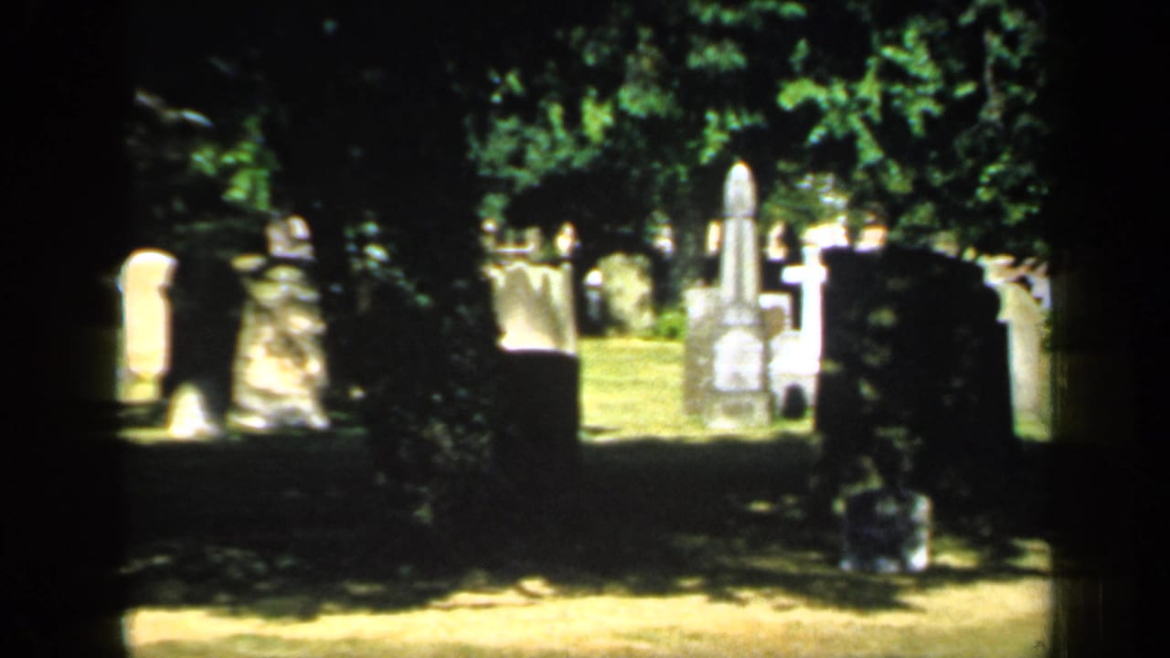 1949 England home-movie frame showing a shaded churchyard with multiple weathered headstones, grass, and sunlight filtering through trees.