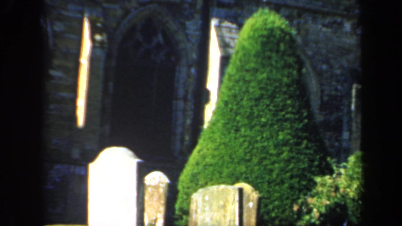 1949 England frame with pale and weathered headstones in front of a clipped evergreen and an old stone church wall with arched openings.
