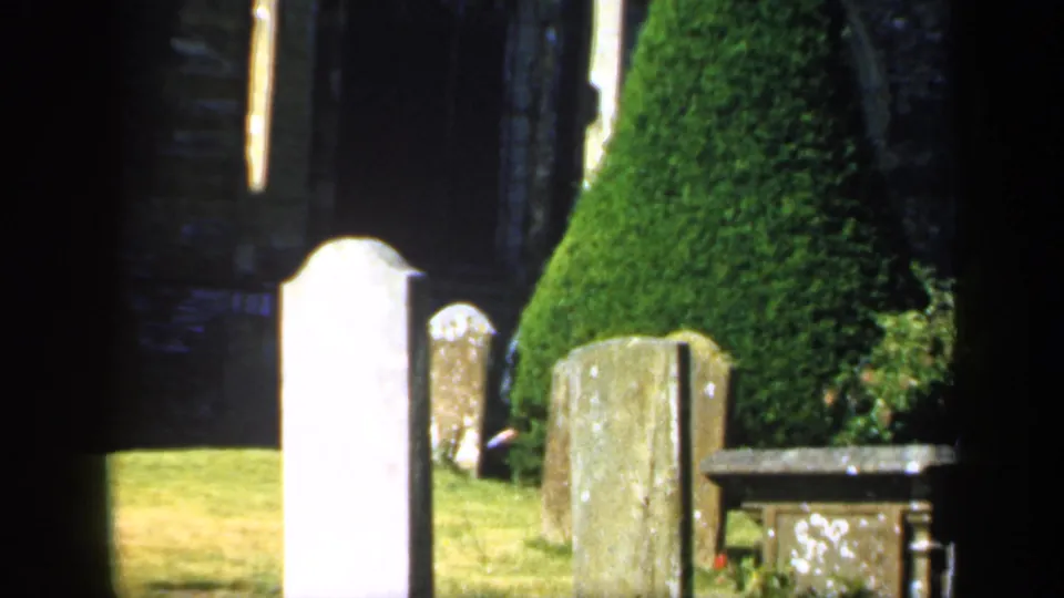 1949 England home-movie frame showing a closer view of churchyard markers, clipped evergreen topiary, grass, and a low stone tomb or monument.