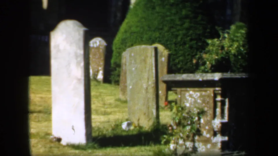 1949 England frame with sunlit headstones, dry grass, a clipped evergreen, and a low stone monument at the edge of the churchyard view.