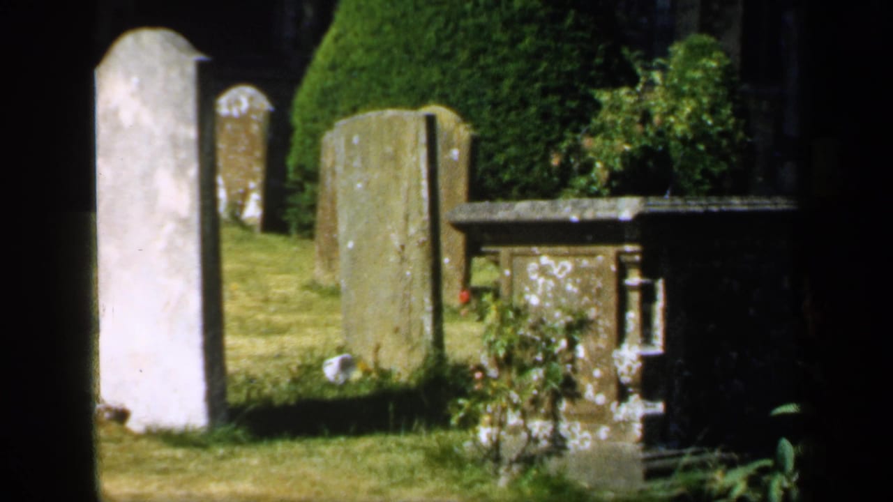 1949 England home-movie frame showing a sunlit churchyard with weathered gravestones, trimmed hedges, grass, and a stone memorial table.