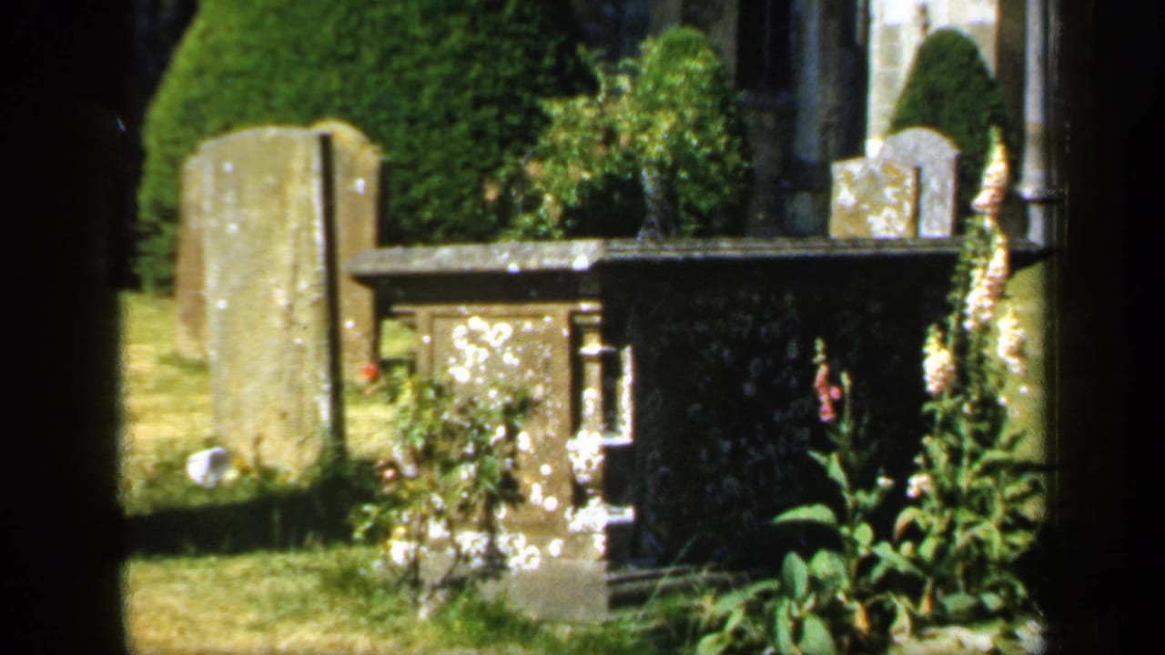 1949 England cemetery view with weathered gravestones, a lichen-marked stone memorial, clipped greenery, flowers, and church stonework behind.