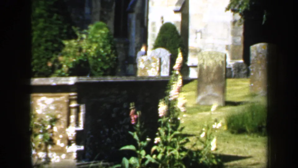 1949 England home-movie frame of a churchyard with gravestones, tall foxgloves, hedges, grass, and a distant adult visitor near the church wall.