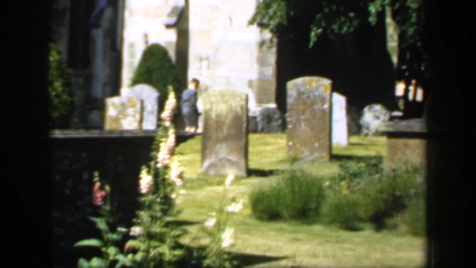 1949 England churchyard frame showing gravestones on a green lawn, foxgloves in the foreground, trimmed hedges, church stonework, and a distant suited man.