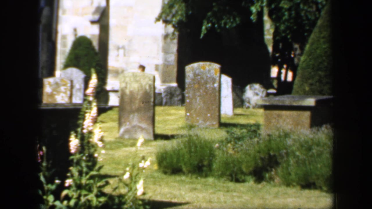 English cemetery with weathered gravestones, grass, topiary, and pale stone building beyond.