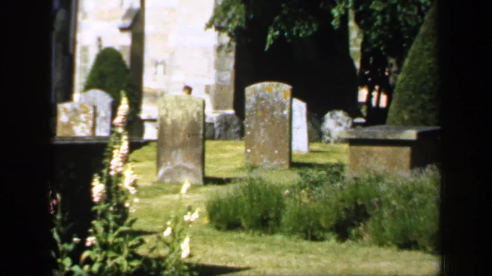 1949 England 8mm frame showing a cemetery lawn with aged gravestones, clipped greenery, trees, and pale masonry in the background.