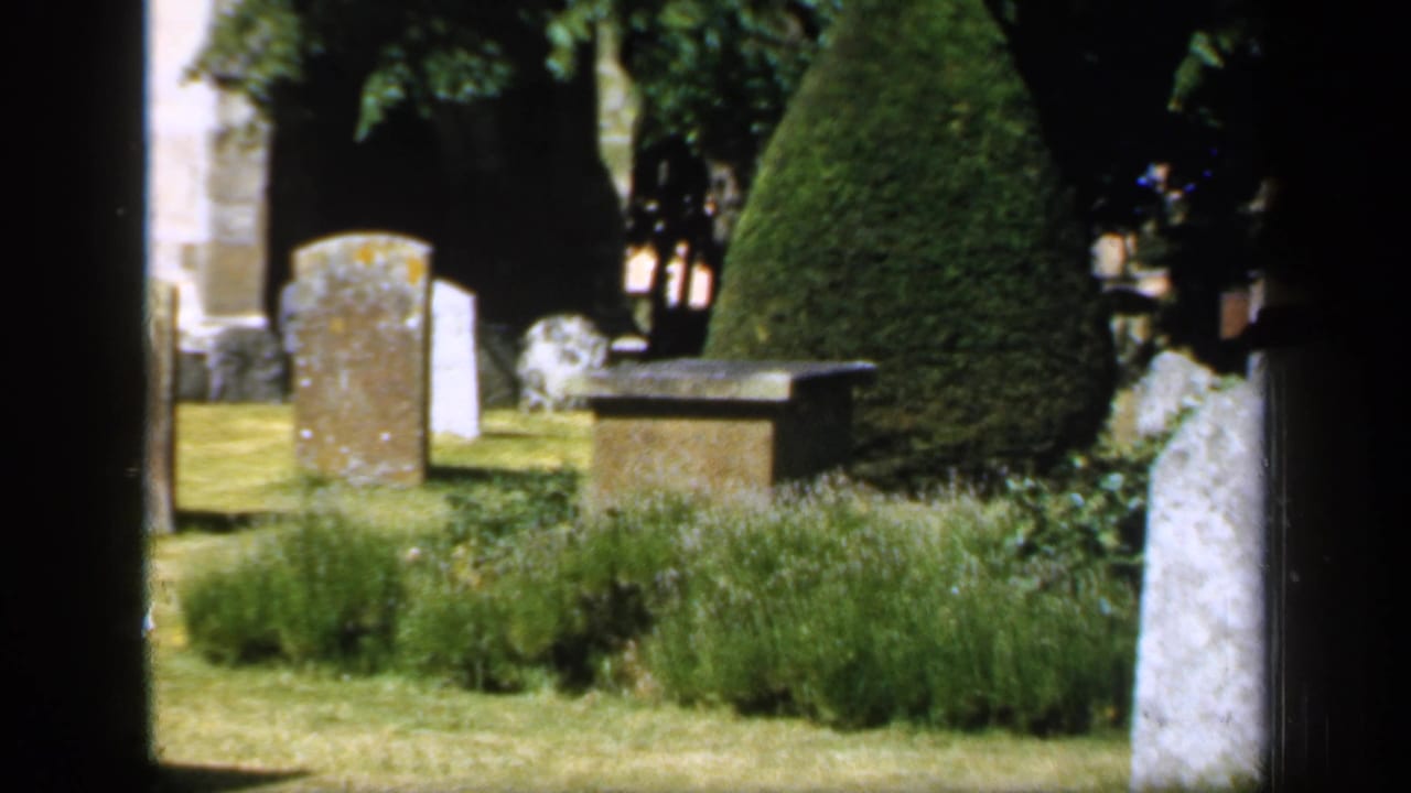 1949 England 8mm frame of a churchyard cemetery with multiple stone markers, clipped topiary, shrub beds, and a low stone structure.
