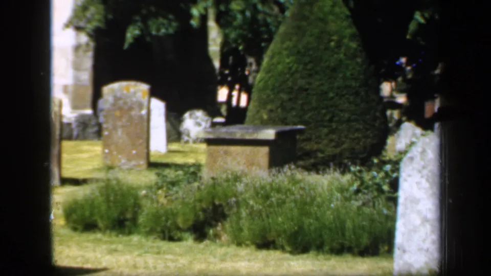 1949 England 8mm frame of a churchyard cemetery with multiple stone markers, clipped topiary, shrub beds, and a low stone structure.