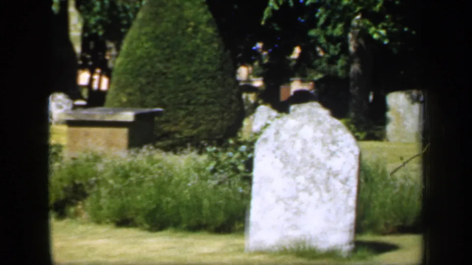 1949 England 8mm frame centered on a pale grave marker, with grass, shrubs, topiary, and additional cemetery stones behind it.