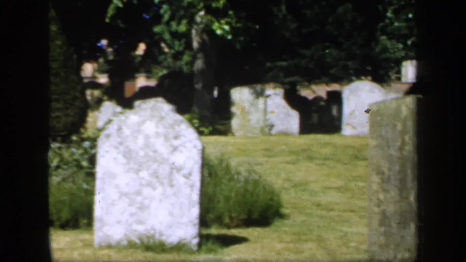 1949 England 8mm frame showing a foreground cemetery headstone, grassy ground, shrubs, and several pale markers under tree shade.