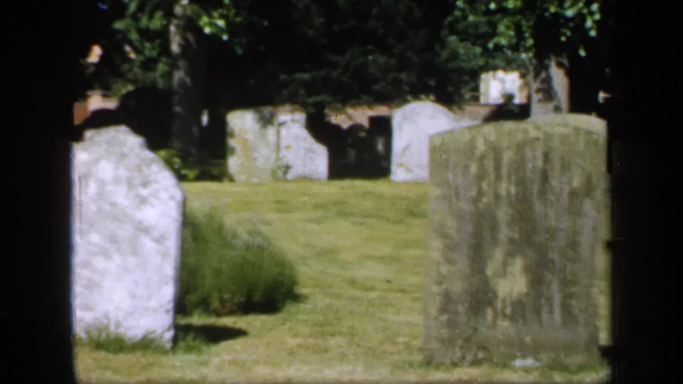 1949 England 8mm frame of weathered upright gravestones in a quiet grassy cemetery with mature trees and shaded edges.