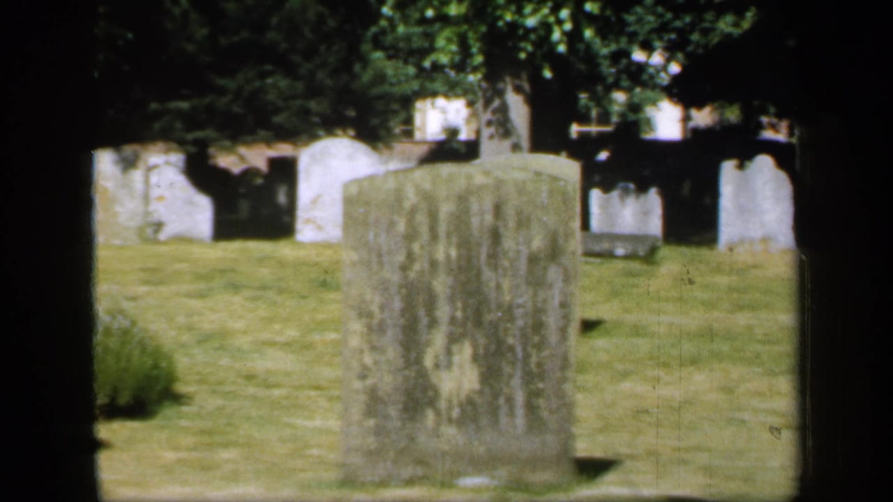 A large weathered headstone stands on grass with other graves behind it.