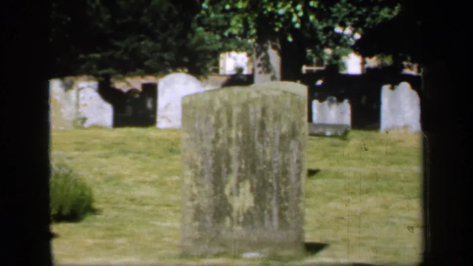 1949 England 8mm view of a worn headstone in the foreground, with more grave markers and trees across a sunlit cemetery lawn.