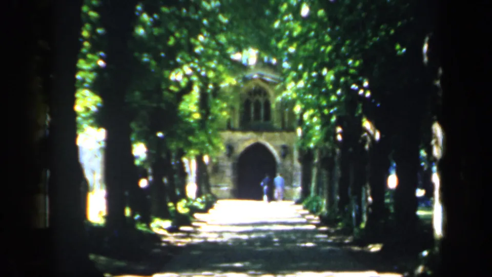 1949 England 8mm frame showing a shaded tree-lined path toward a historic stone archway, with distant visitors visible.