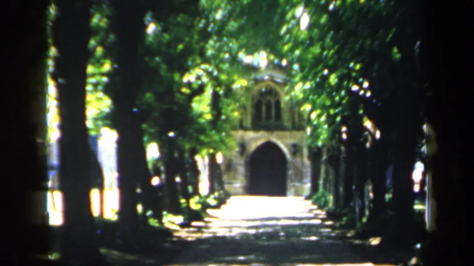 1949 England 8mm frame of a leafy path leading to a distant stone archway, with heavy shade and bright sun patches.