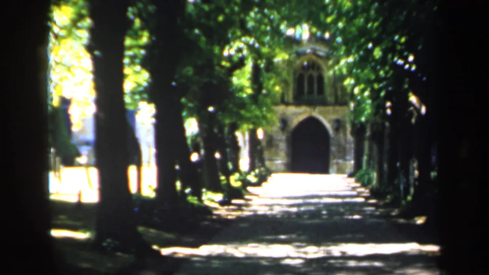 1949 England 8mm frame showing a shaded tree-lined path with bright dappled sunlight and a distant historic stone facade.