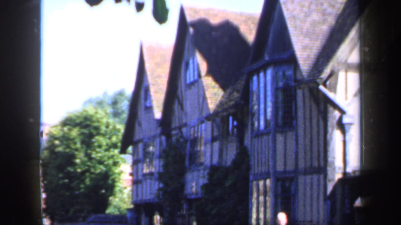 1949 England home-movie frame showing traditional timber-framed village houses partly obscured by a dark post or tree and leaves.