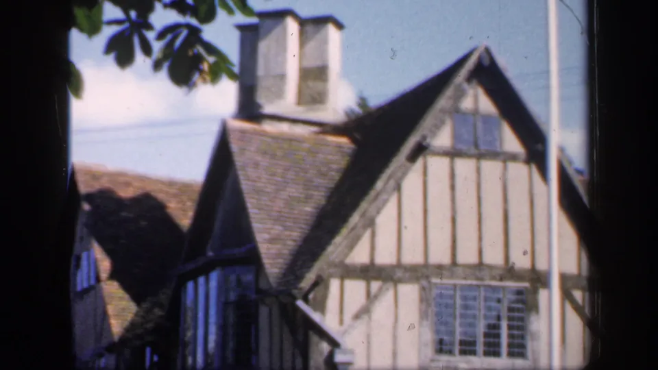 A traditional half-timbered house in England, filmed in 1949, shows a gabled facade, leaded window, tiled roof, and chimney.