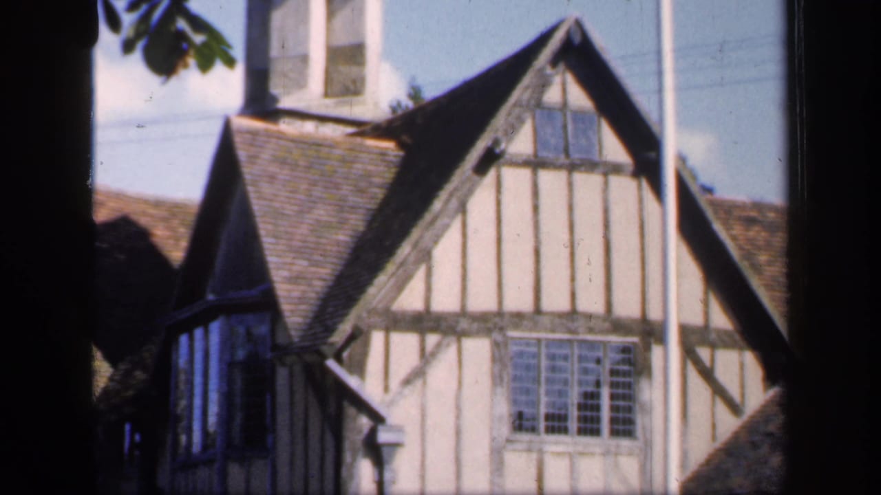 1949 England home-movie frame showing a traditional timber-framed house with cream plaster, dark beams, steep roof, and multi-pane window.