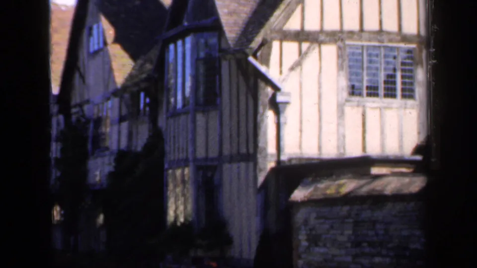 1949 England home-movie view of a half-timbered building facade with a leaded window, shaded wall, and low stone boundary.